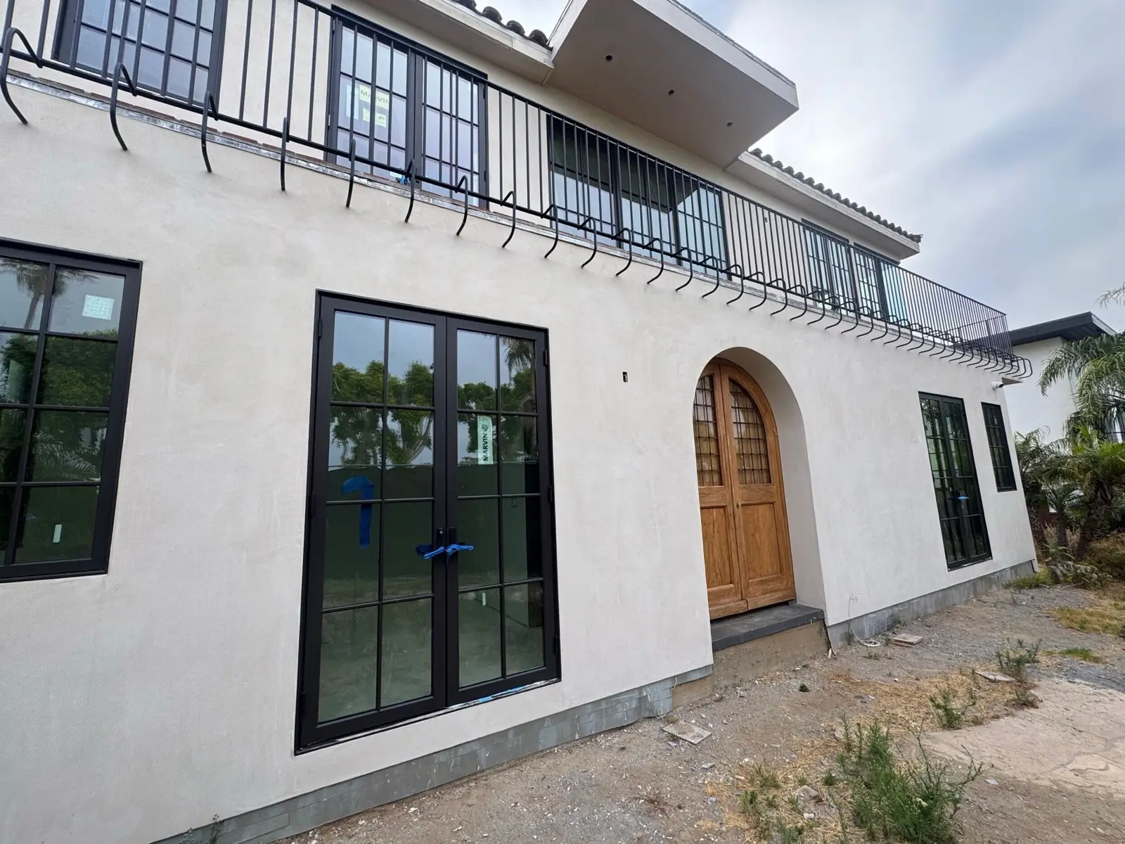 A Farnsworth Builders home under construction — stucco exterior with black steel-framed windows and an arched timber front door, before final grading.